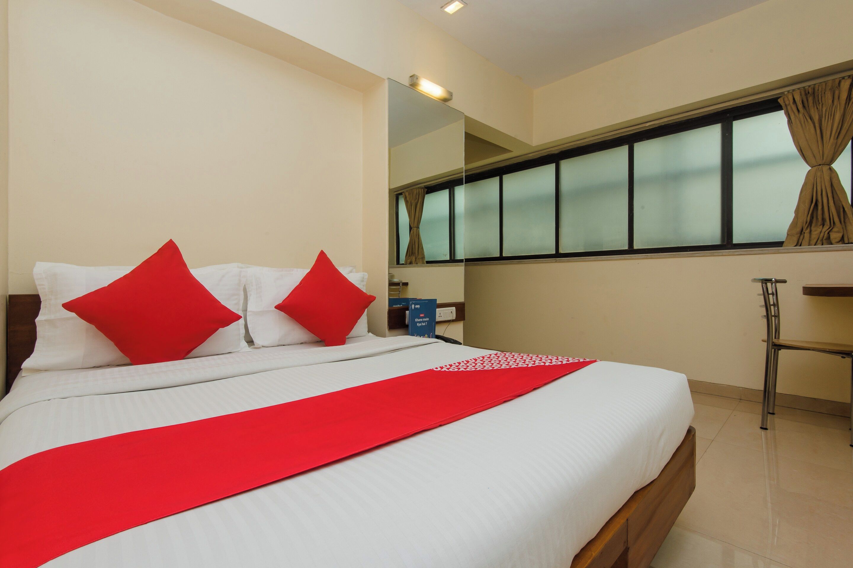 Quarto de hotel com cama, manta vermelha, mesa de cabeceira, espelho e janela com cortinas.