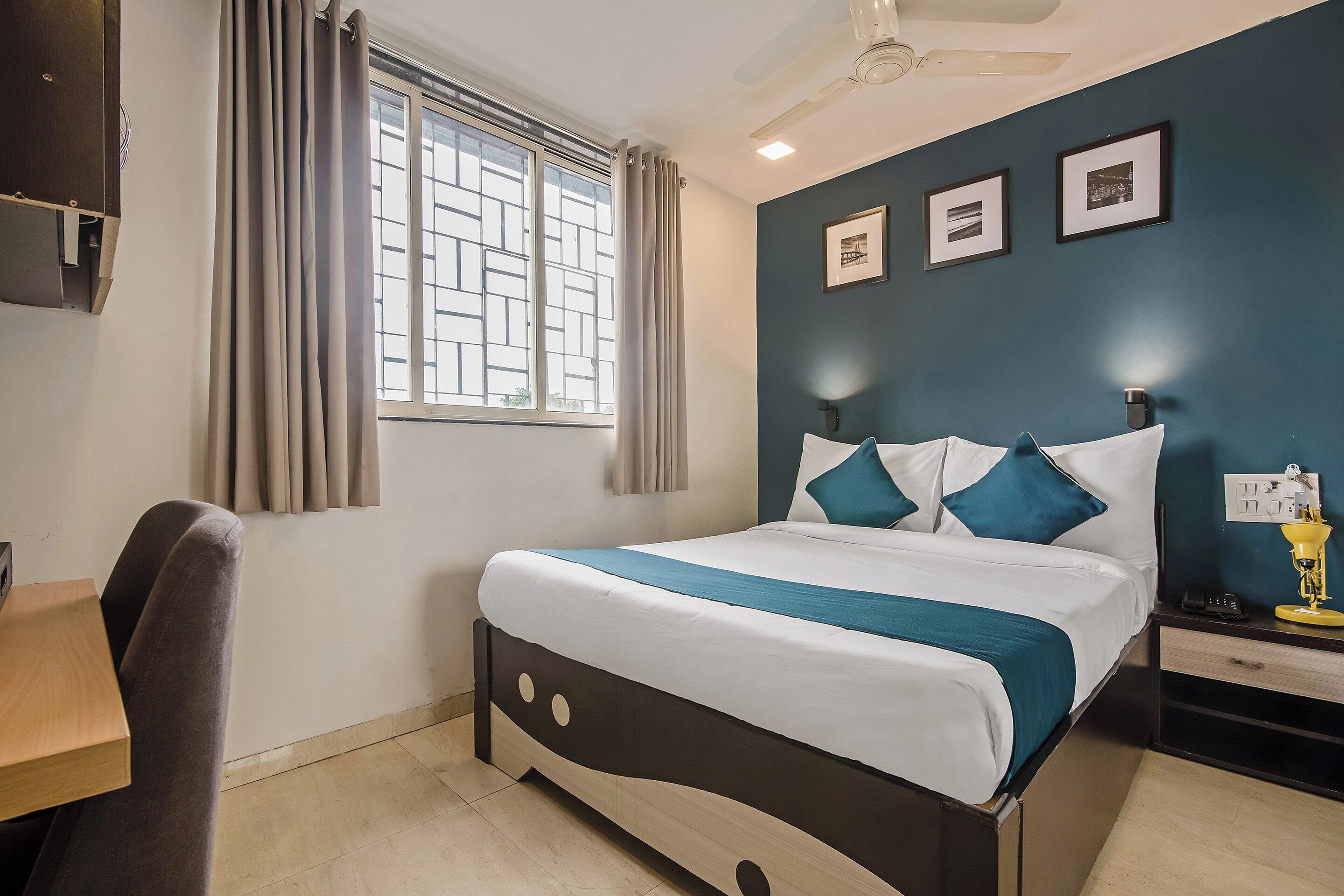 A hotel room with a bed, a desk, a chair, a window with curtains, a ceiling fan, and wall-mounted lights.