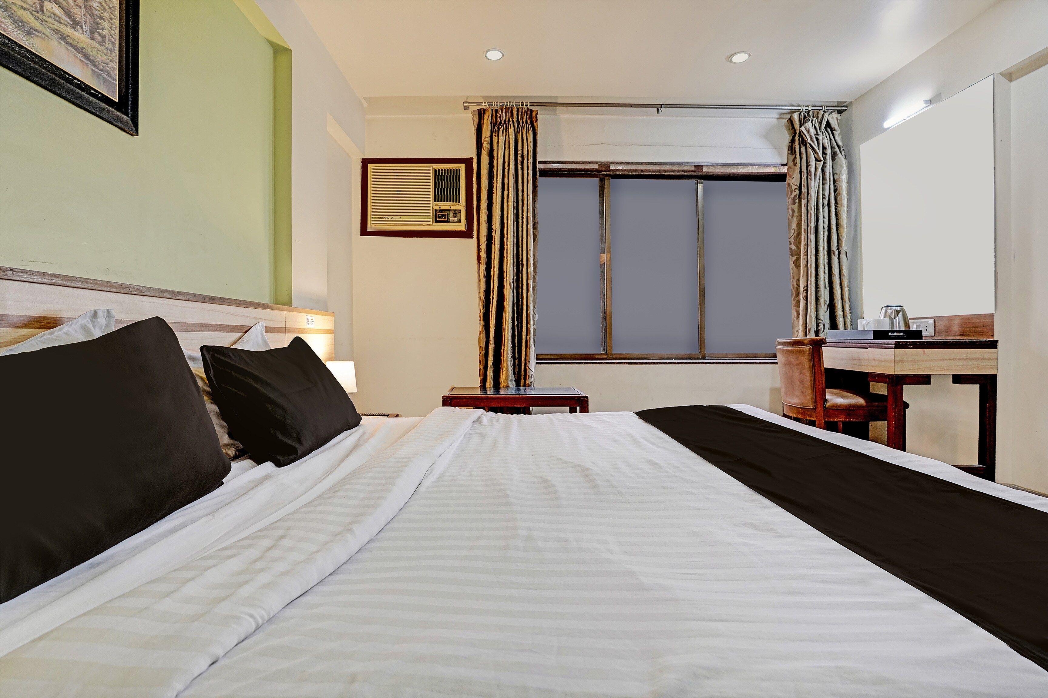 Una habitación de hotel con cama, escritorio, silla, ventana con cortinas e imagen en la pared.