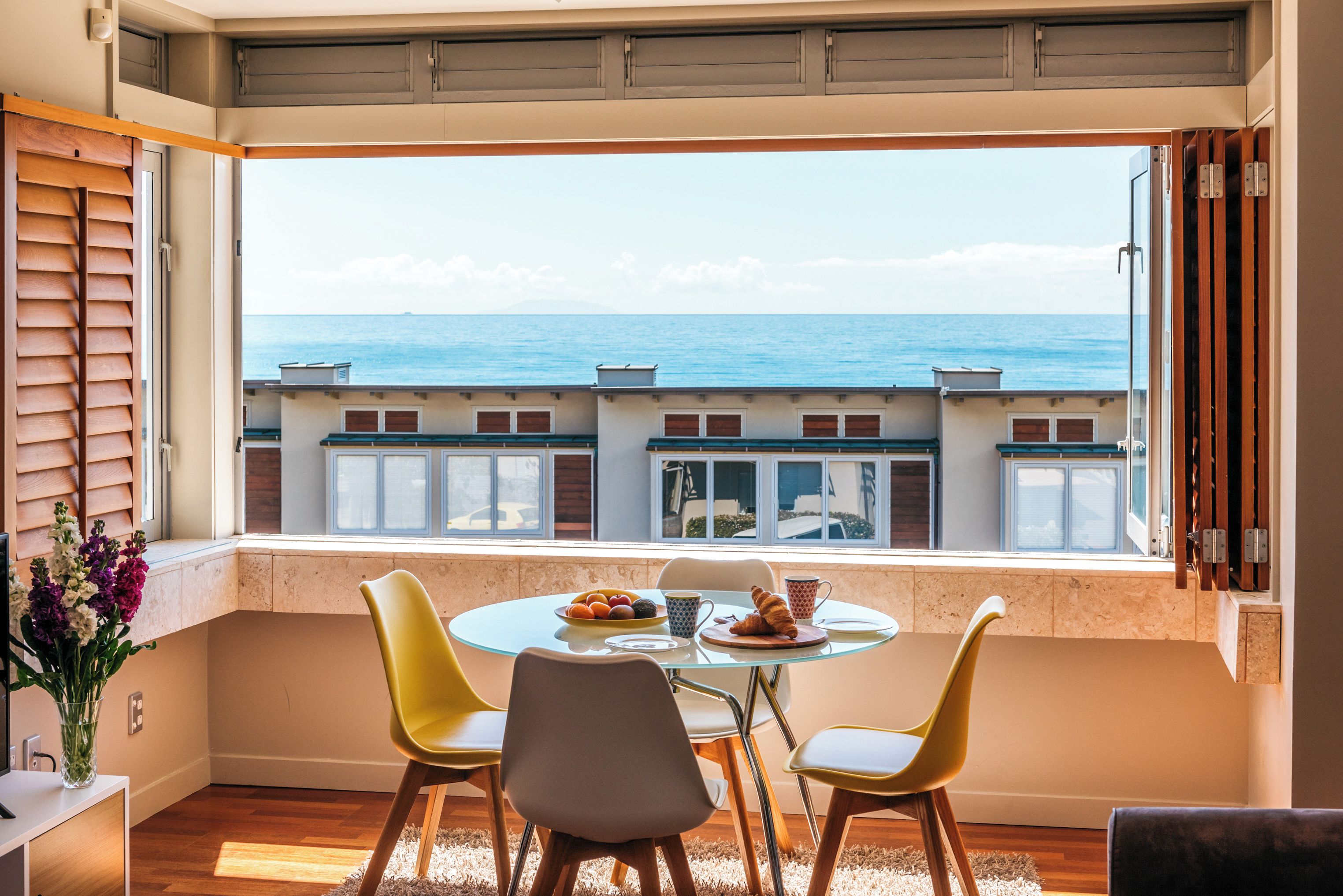 Un comedor con mesa y sillas, vistas al mar y un balcón con contraventanas de madera.