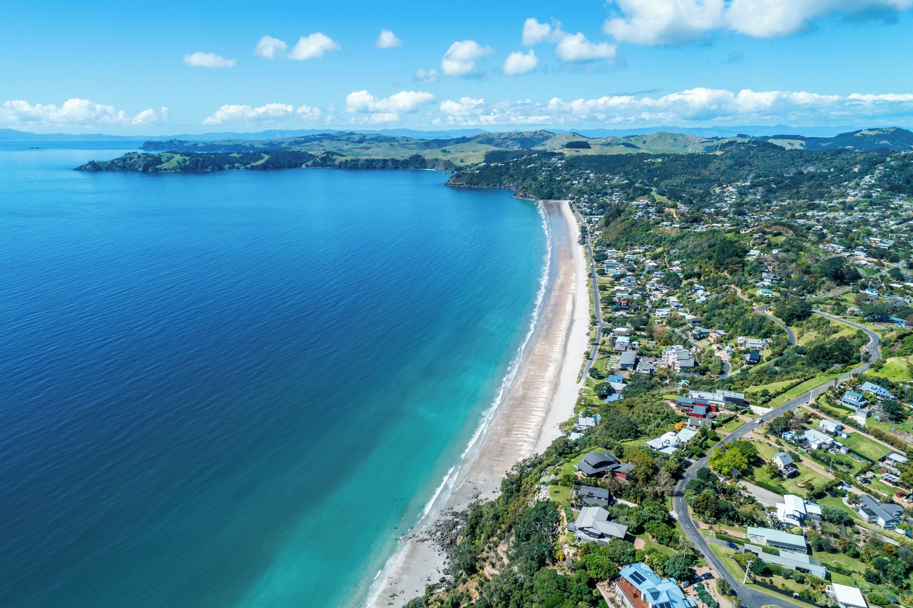 Vista aérea de un pueblo costero con una playa extensa, zonas residenciales y colinas onduladas al fondo.