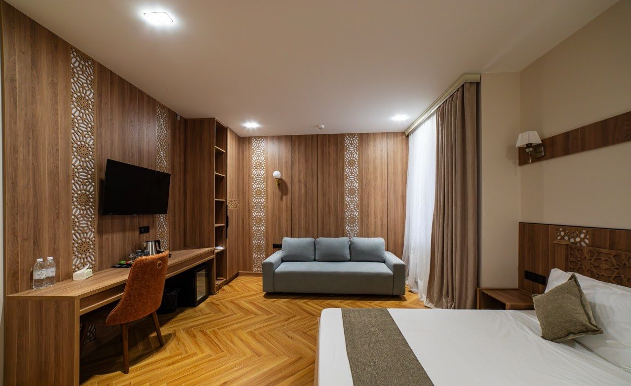 Habitación de hotel con cama, sofá, escritorio con silla, televisión y ventana con cortinas.