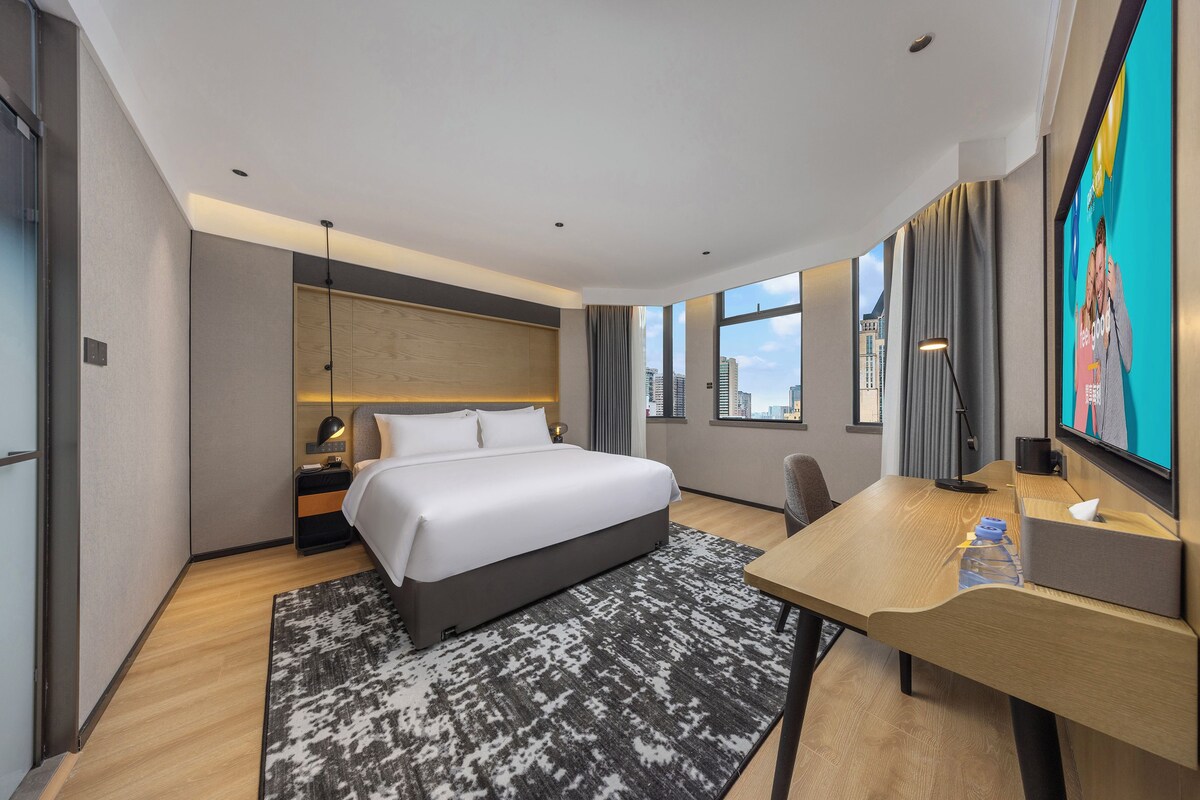 Een hotelkamer met een bed, bureau, stoel en een groot raam met uitzicht op de stad.