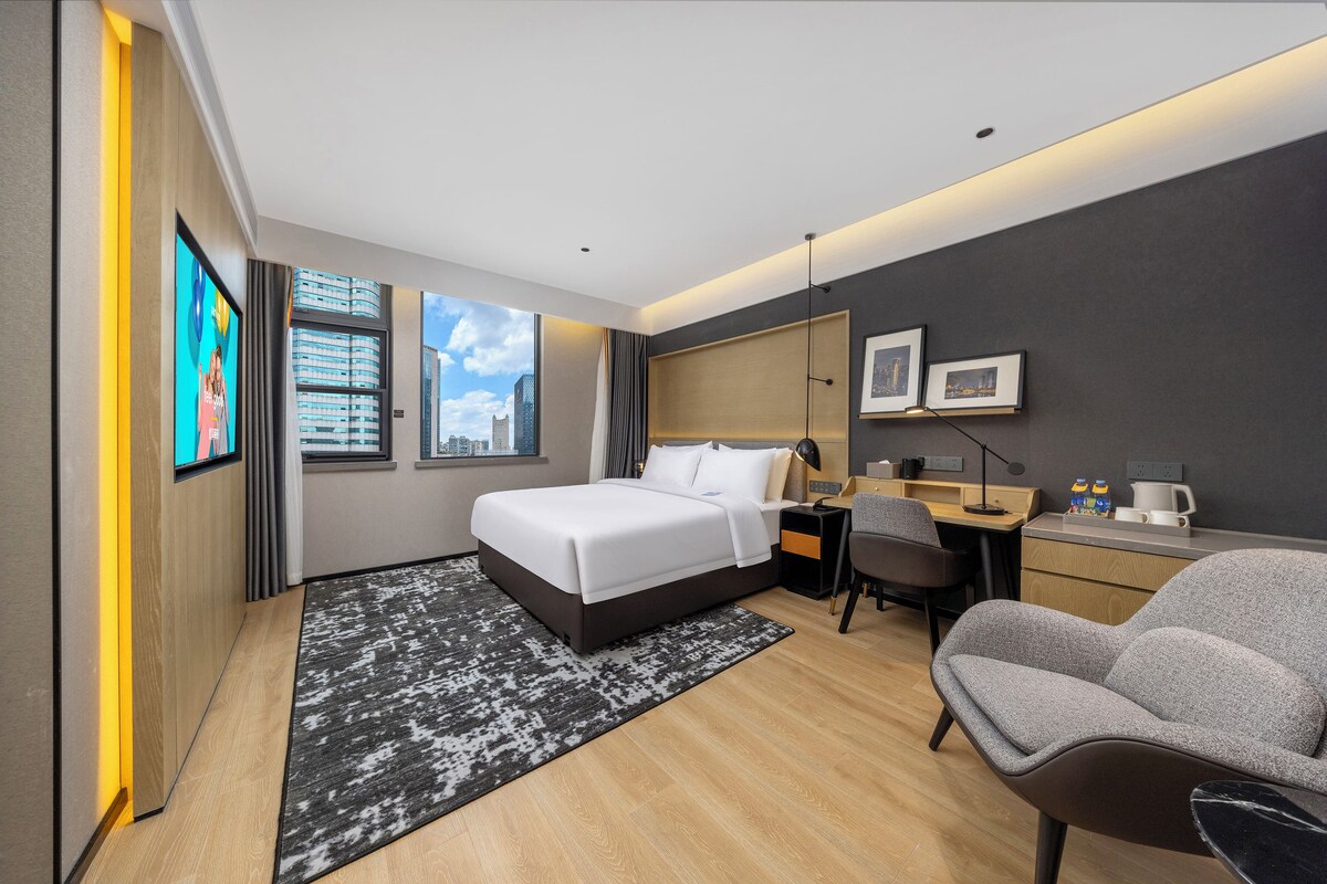 Een hotelkamer met een bed, bureau, stoel en grote ramen met uitzicht op de stad.
