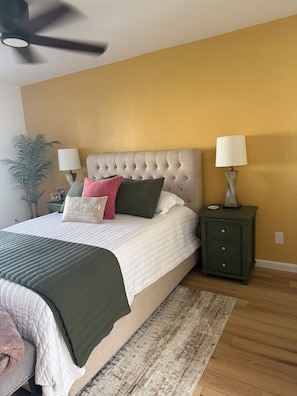 A bedroom with a tufted headboard, a bed with multiple pillows, two bedside lamps, a nightstand, a potted plant, and a wooden floor. - “Cactus Casita” 2-bedroom condo in Westbrook Village (Peoria)