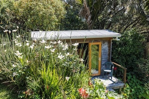 A small wooden structure with a metal roof, surrounded by lush greenery and white flowers. - Palm Beach Panorama - A readers dream home with views of Palm Beach (Waiheke Island)