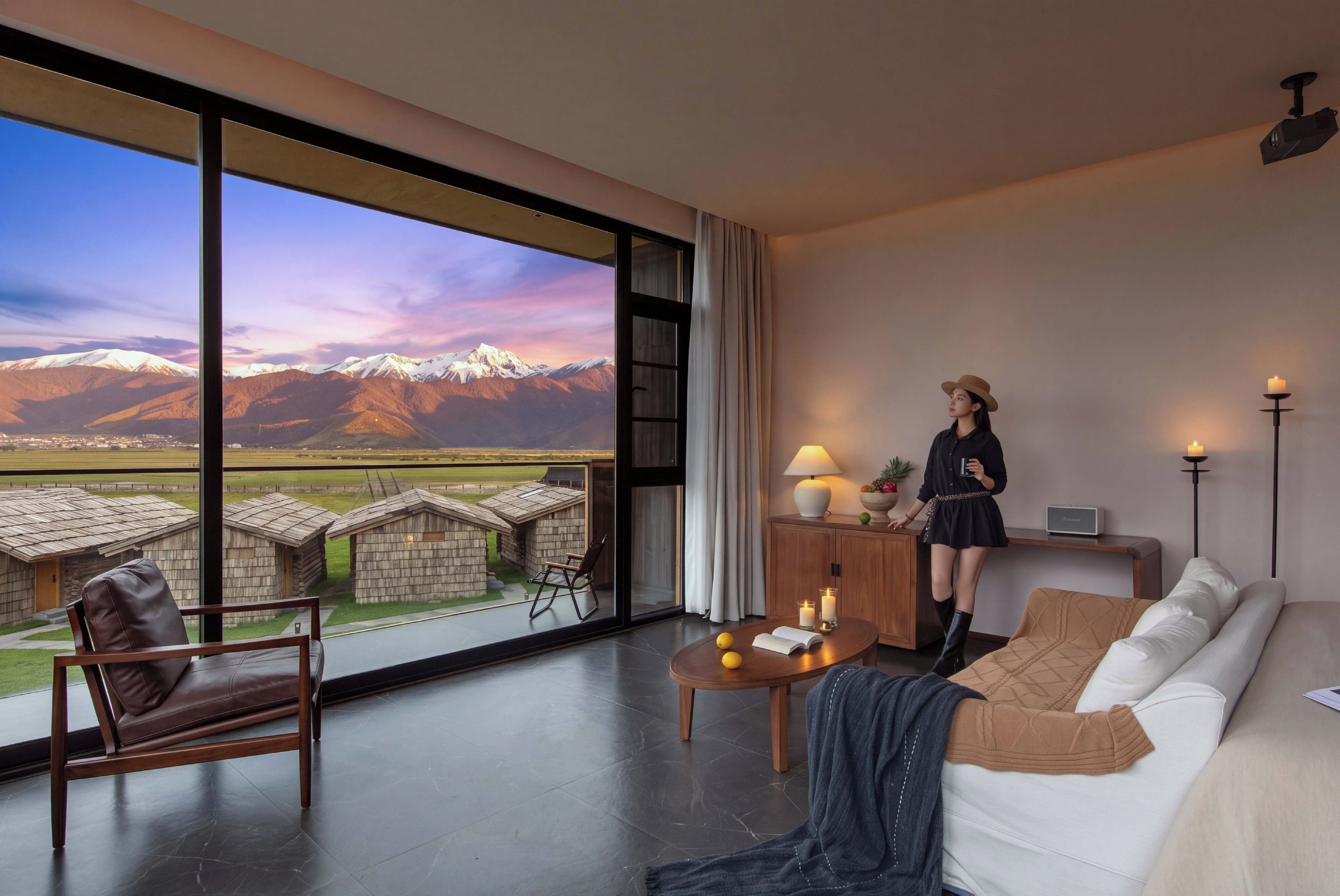 A modern hotel room with a large window offering a mountain view, a sofa, a chair, a small table, and a lamp.