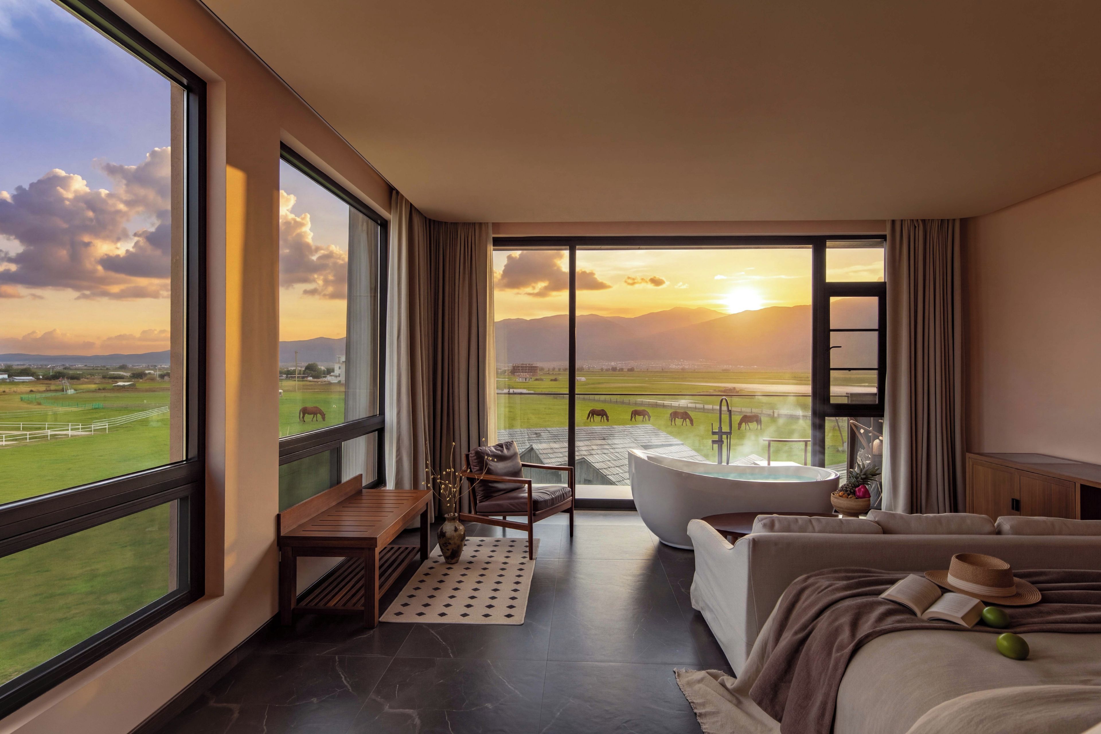 A modern hotel room with a large window offering a view of a green landscape and mountains.
