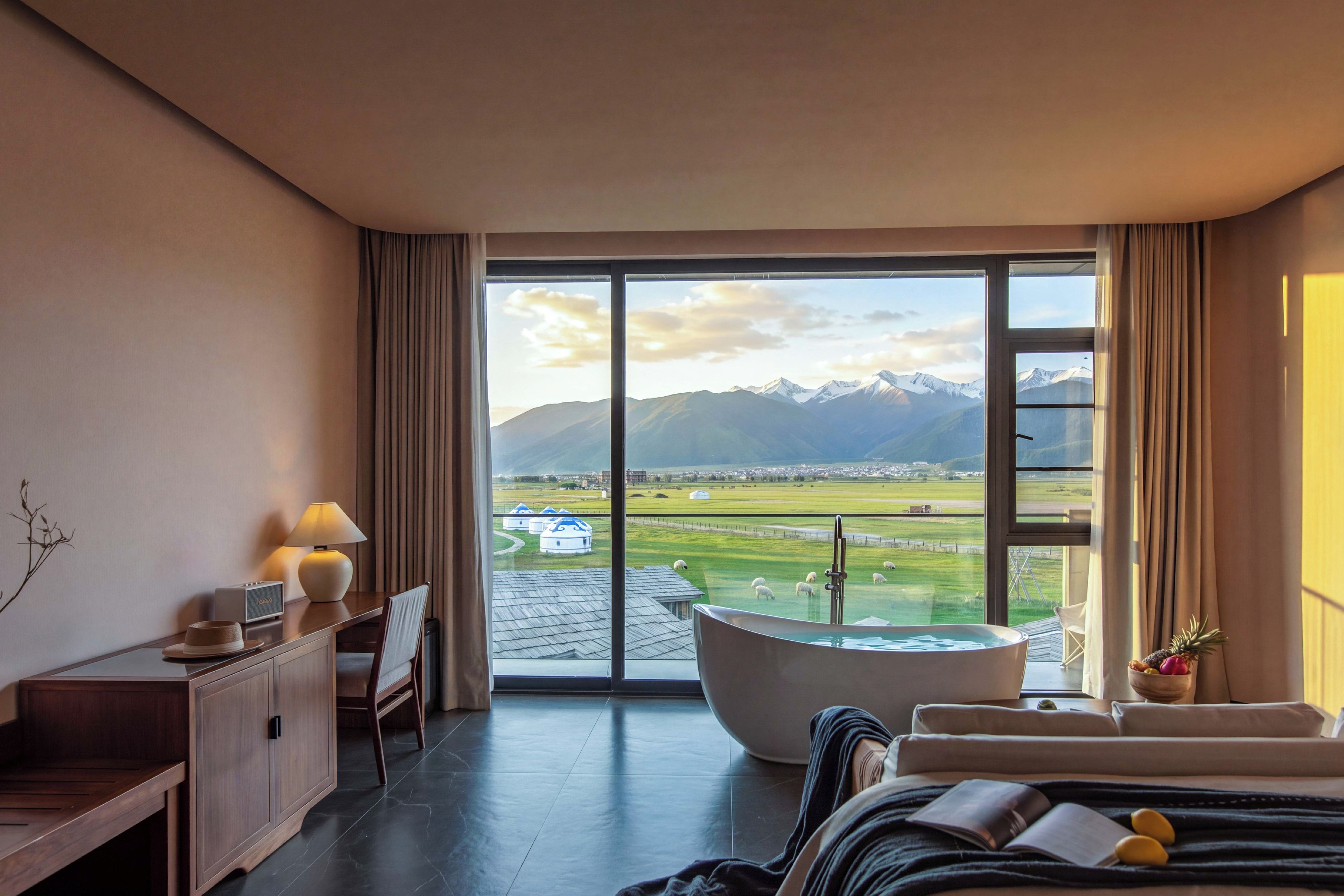 A hotel room with a large window, a bathtub, a desk with a lamp, and a bed.