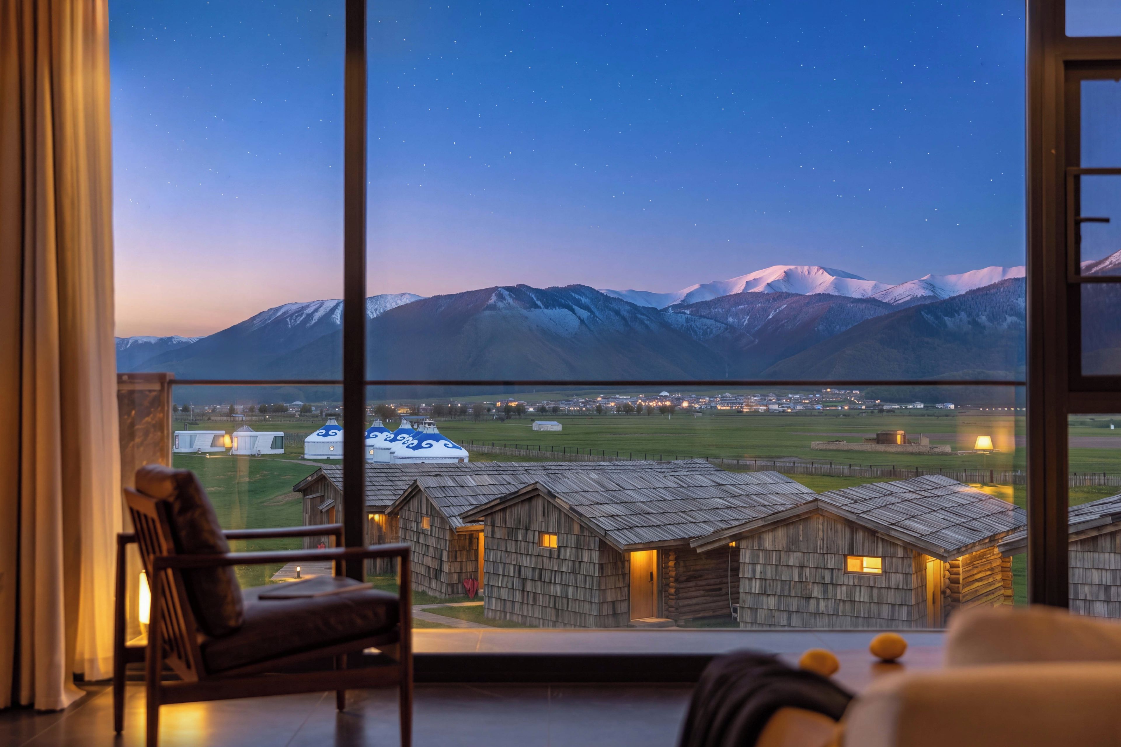 A room with a large window overlooking a mountainous landscape and yurts.