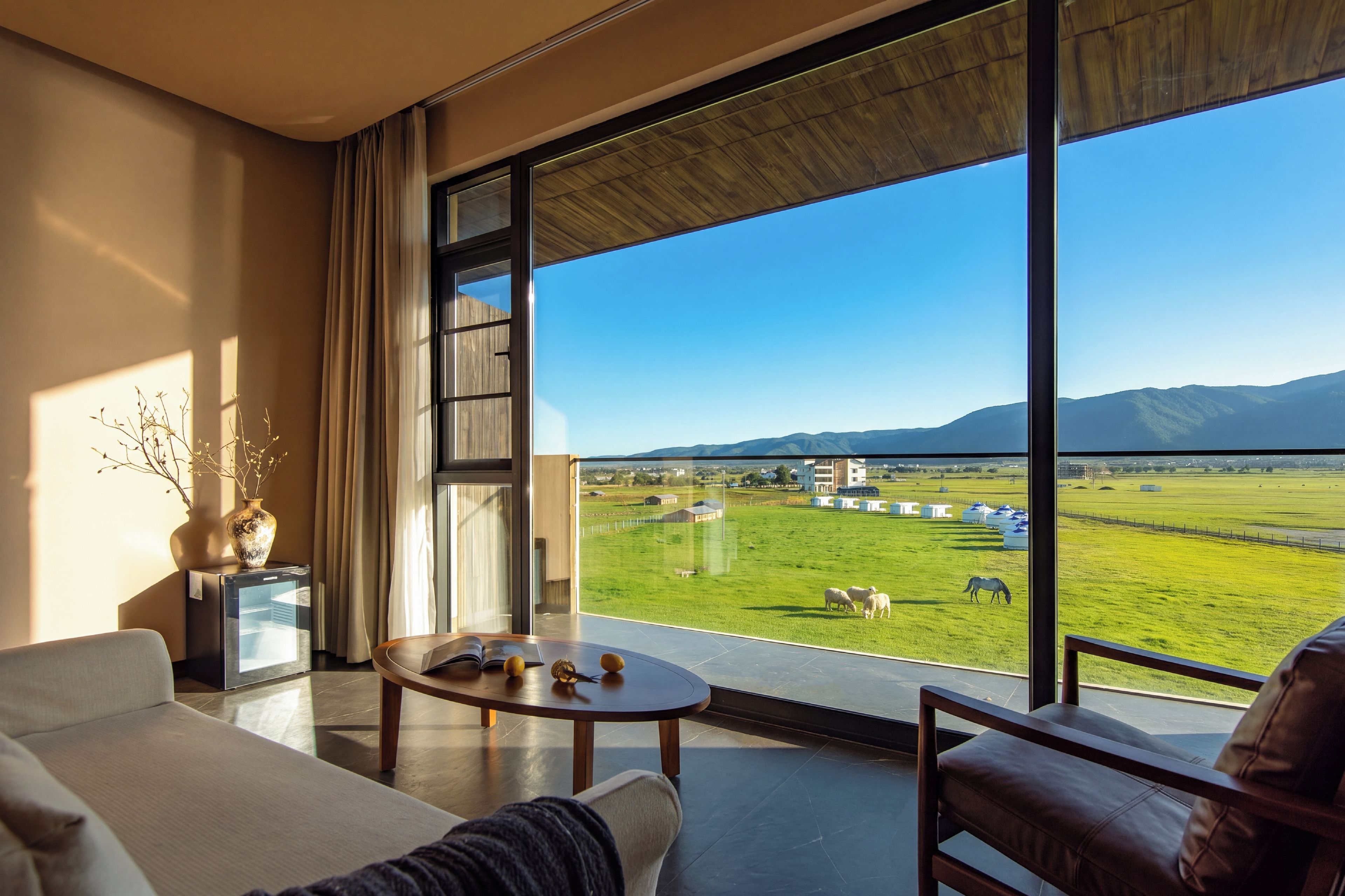 A modern room with a large window, a sofa, a coffee table, and a view of a green field with animals.