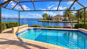 A swimming pool with a clear view of the water, a covered area with a roof, and a distant view of a body of water and buildings. - Stunning 4-Bedroom Riverfront Retreat with Private Hot Tub & Heated Pool! (Cape Coral)