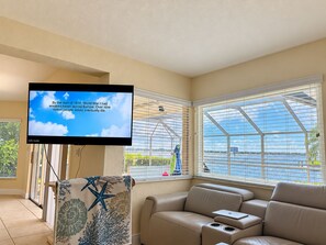 A living room with a television displaying a historical message, a sofa, and a view through a large window. - Stunning 4-Bedroom Riverfront Retreat with Private Hot Tub & Heated Pool! (Cape Coral)