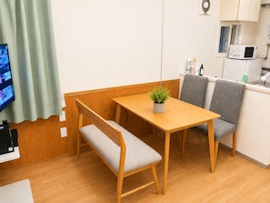 Ein Zimmer mit Holztisch, zwei grauen Stühlen, einer Bank, einem kleinen Topfpflanzen und einem Fernseher, sowie einer Küchenzeile mit einer Mikrowelle.