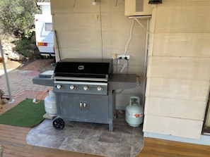 A barbecue grill with a gas tank and a propane cylinder, a wooden deck, and a vehicle in the background. - Shack 1956 (Younghusband)