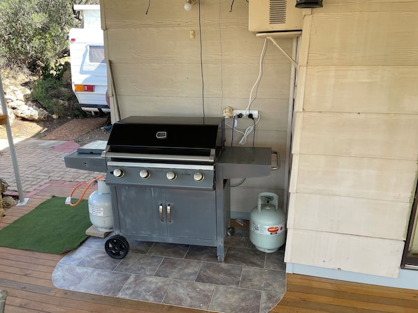 A barbecue grill with a gas tank and a propane cylinder, a wooden deck, and a vehicle in the background.