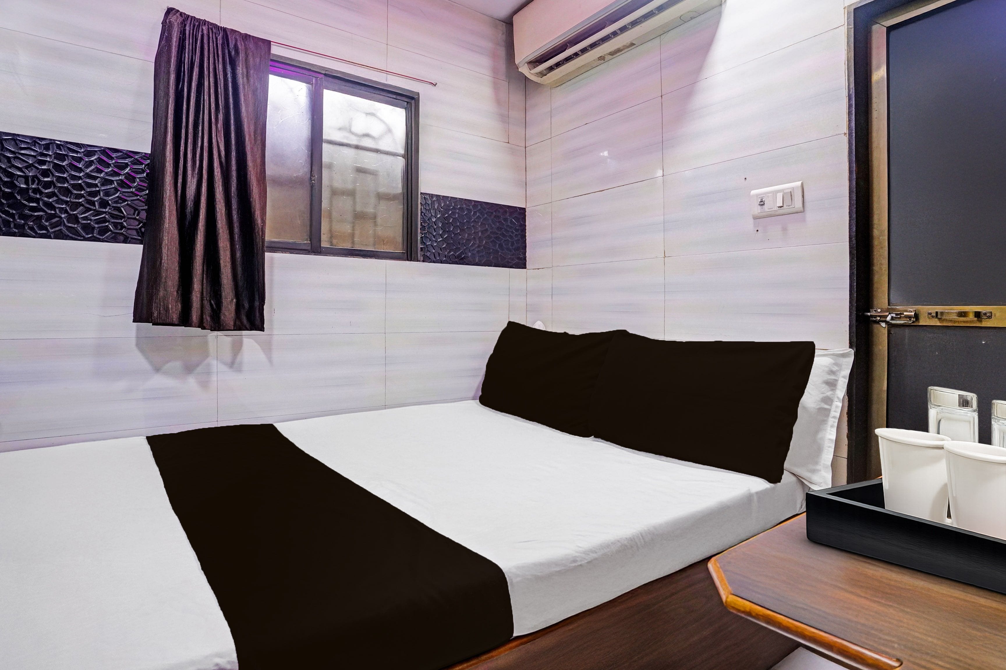 A single bed with a white and black bedspread, a window with a curtain, and a wall-mounted air conditioner.