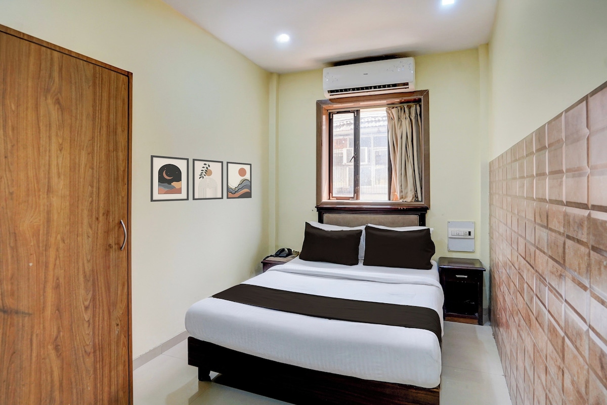 A hotel room with a bed, bedside table, nightstand, wardrobe, air conditioning unit, window with curtains, and wall-mounted artwork.