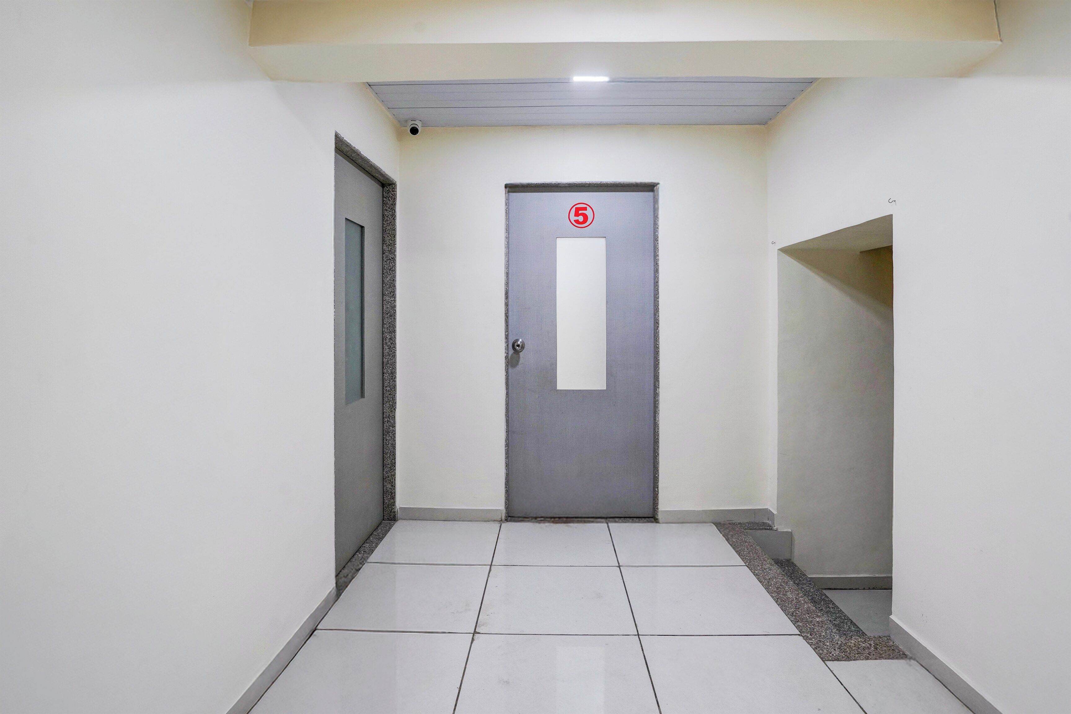 Un pasillo con una puerta numerada, suelo de baldosas y una lámpara en el techo.