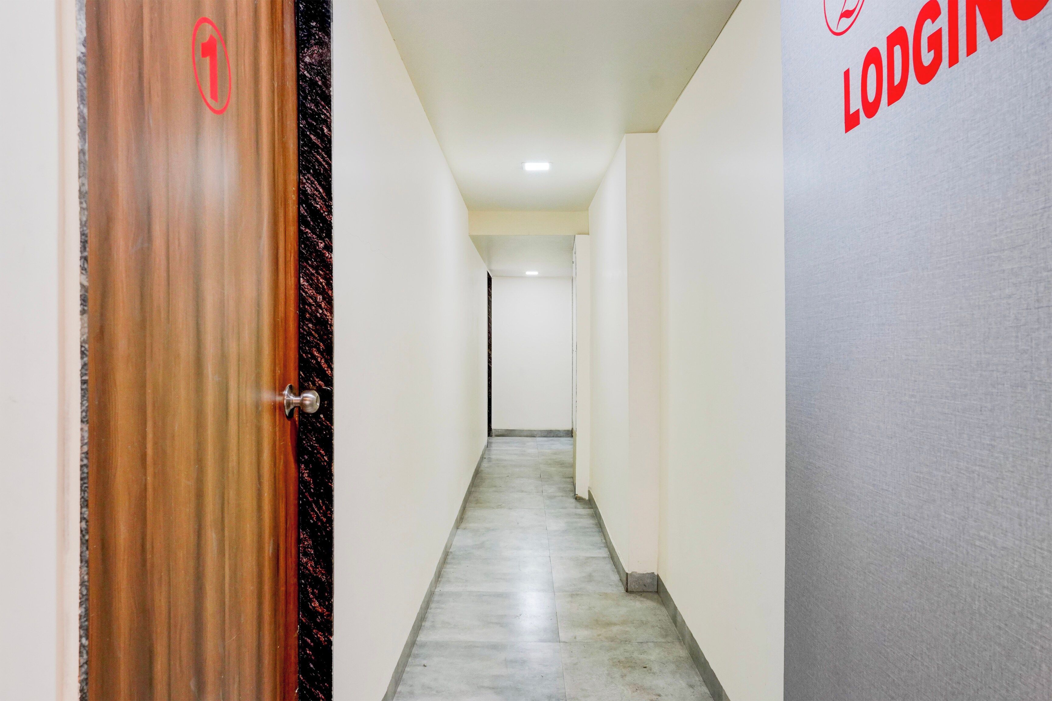 Un pasillo con una puerta de madera marcada con el número 1, una pared gris con la palabra 'LODGING' en rojo y un piso de baldosas.