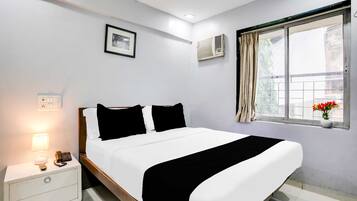 Habitación de hotel con cama, mesita de noche, lámpara, teléfono, equipo de aire acondicionado, ventana con cortinas y un cuadro en la pared.