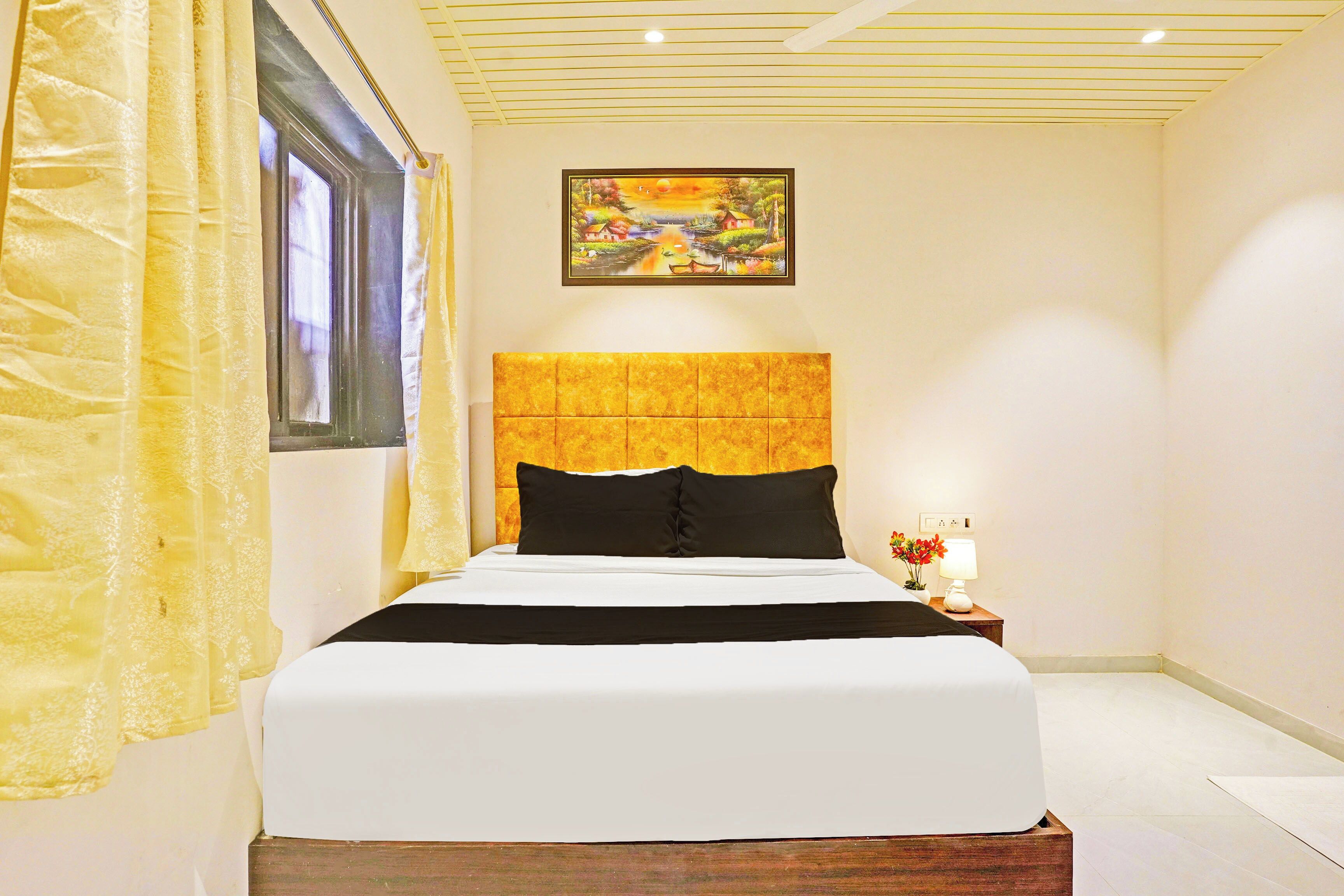 A neatly made bed with a headboard, a window with curtains, a painting on the wall, and a bedside table with a lamp and flowers.