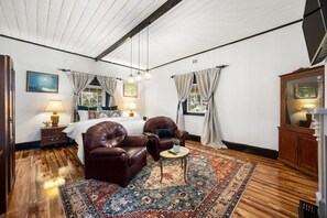 Ein Zimmer mit einem Bett, zwei Ledersesseln, einem runden Tisch, einem gemusterten Teppich und Holzdielen.