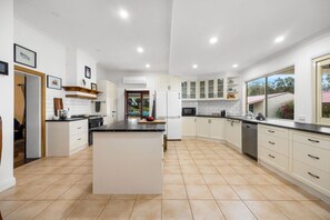 A modern kitchen with white cabinetry, a central island, and a large window providing natural light. - Nori Retreat—Unique Waterfront Heritage on Coorong (Hindmarsh Island)