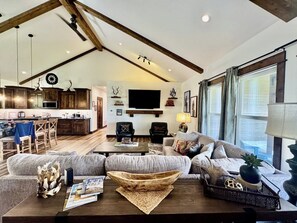 Ein großzügiges Wohnzimmer mit einer großen Couch, einem Couchtisch und einem Fernseher. Im Hintergrund ist eine Küche mit Holzschränken und einem Essbereich zu sehen.