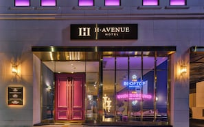 El exterior del HI H AVENUE HOTEL con una entrada destacada, un letrero de neón para el bar y la piscina en la azotea, y un frente de vidrio.