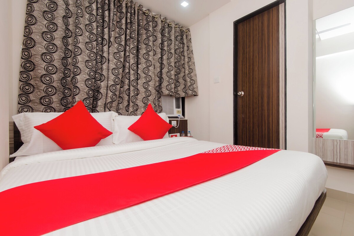 A hotel room with a bed, two red pillows, a red throw, a wooden wardrobe, and a patterned curtain.