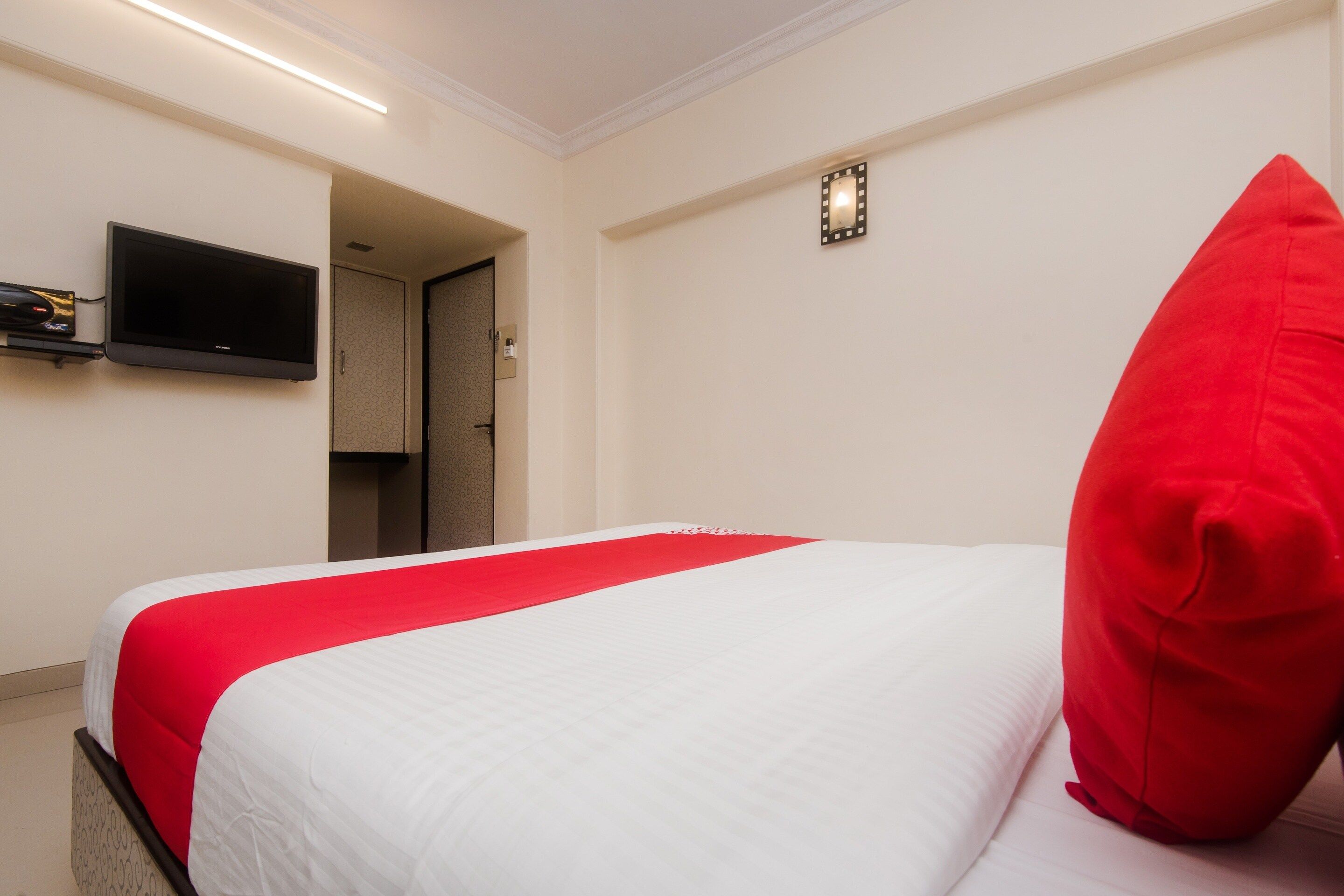 Una habitación de hotel con una cama, un televisor montado en la pared y una almohada roja.