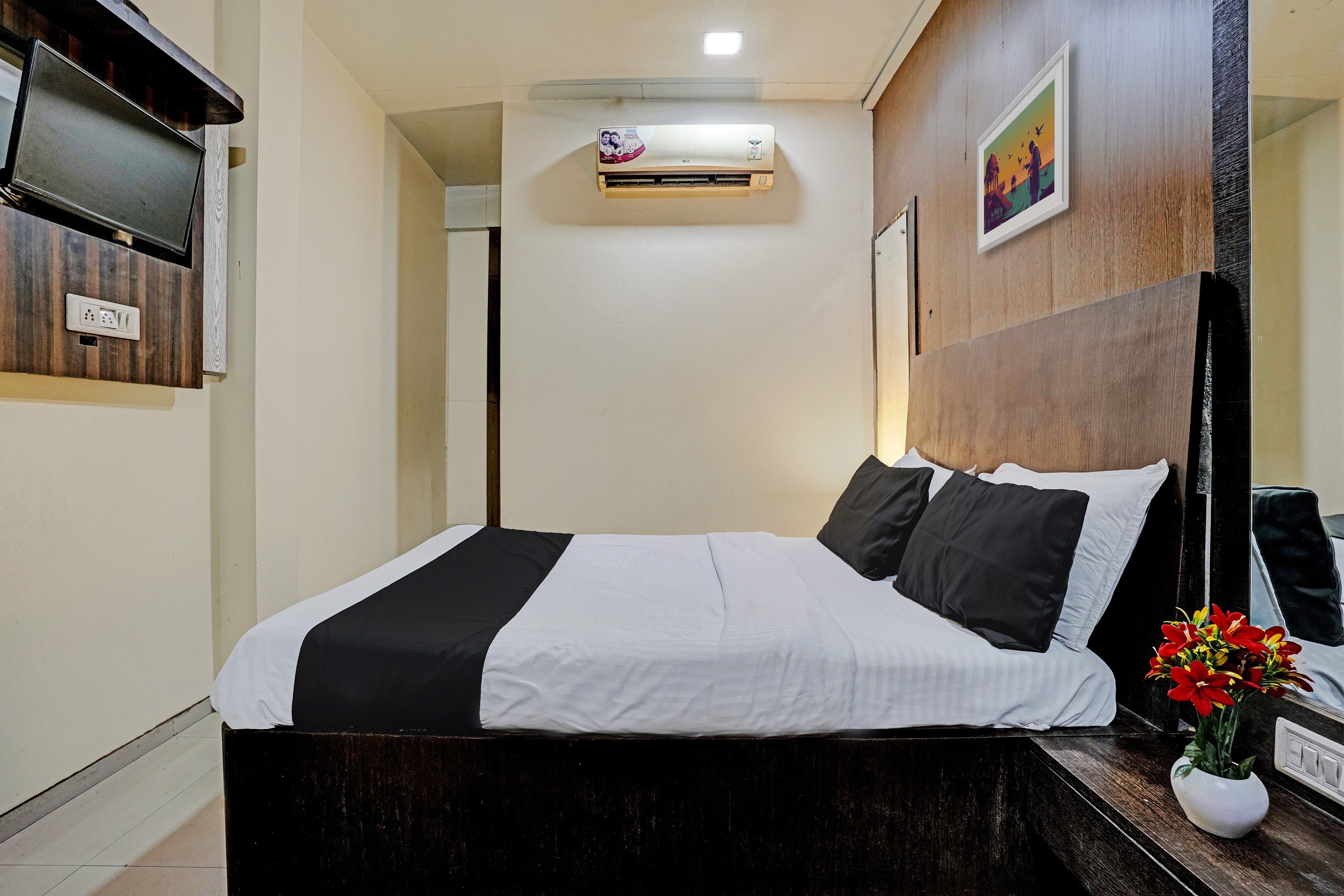A hotel room with a bed, a bedside table with a vase of flowers, an air conditioning unit, and a wall-mounted television.