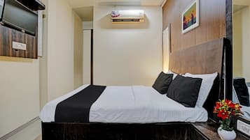 A hotel room with a bed, a bedside table with a vase of flowers, an air conditioning unit, and a wall-mounted television.