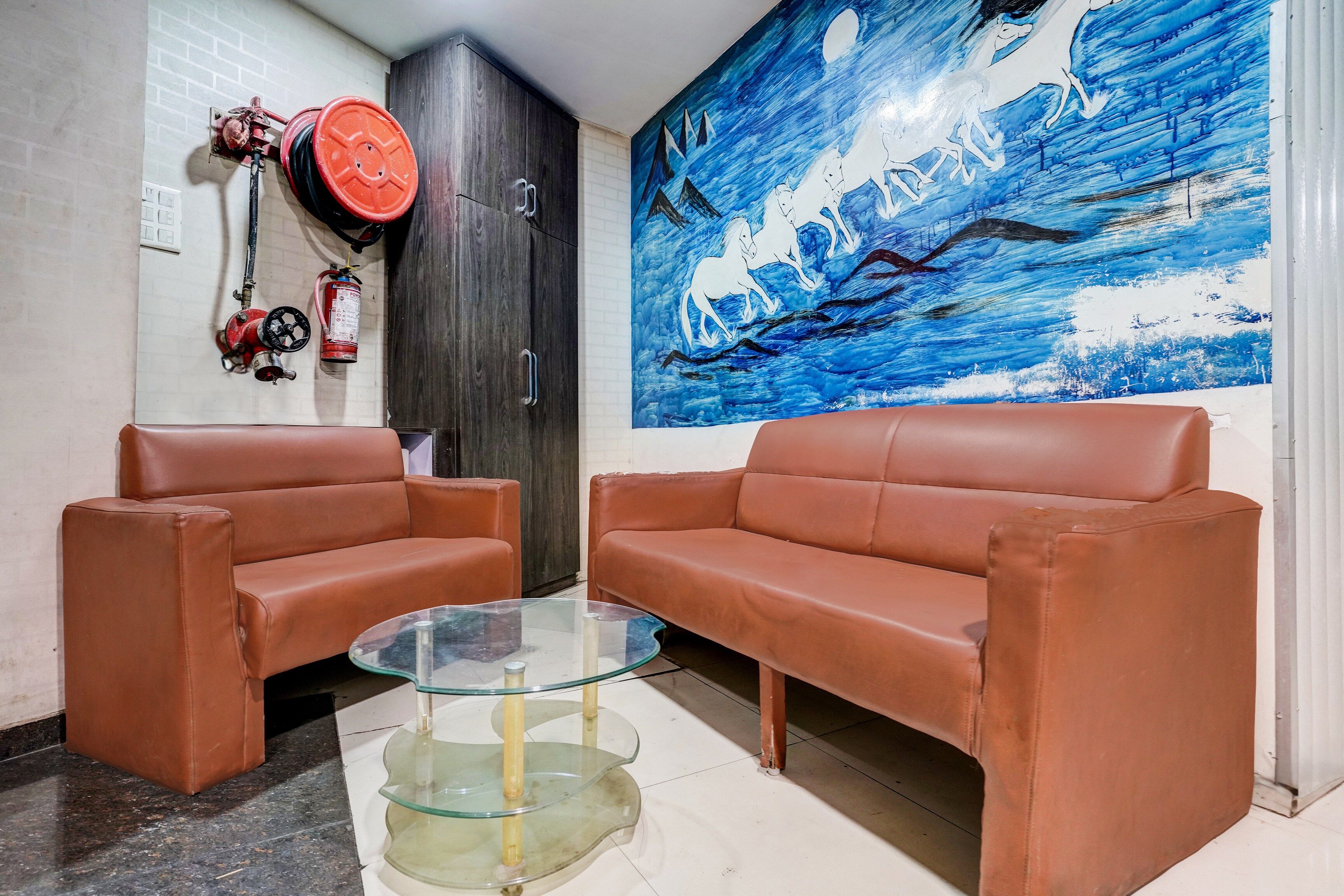 A room with two brown leather sofas, a glass coffee table, a dark wooden wardrobe, a fire extinguisher, and a blue abstract painting on the wall.