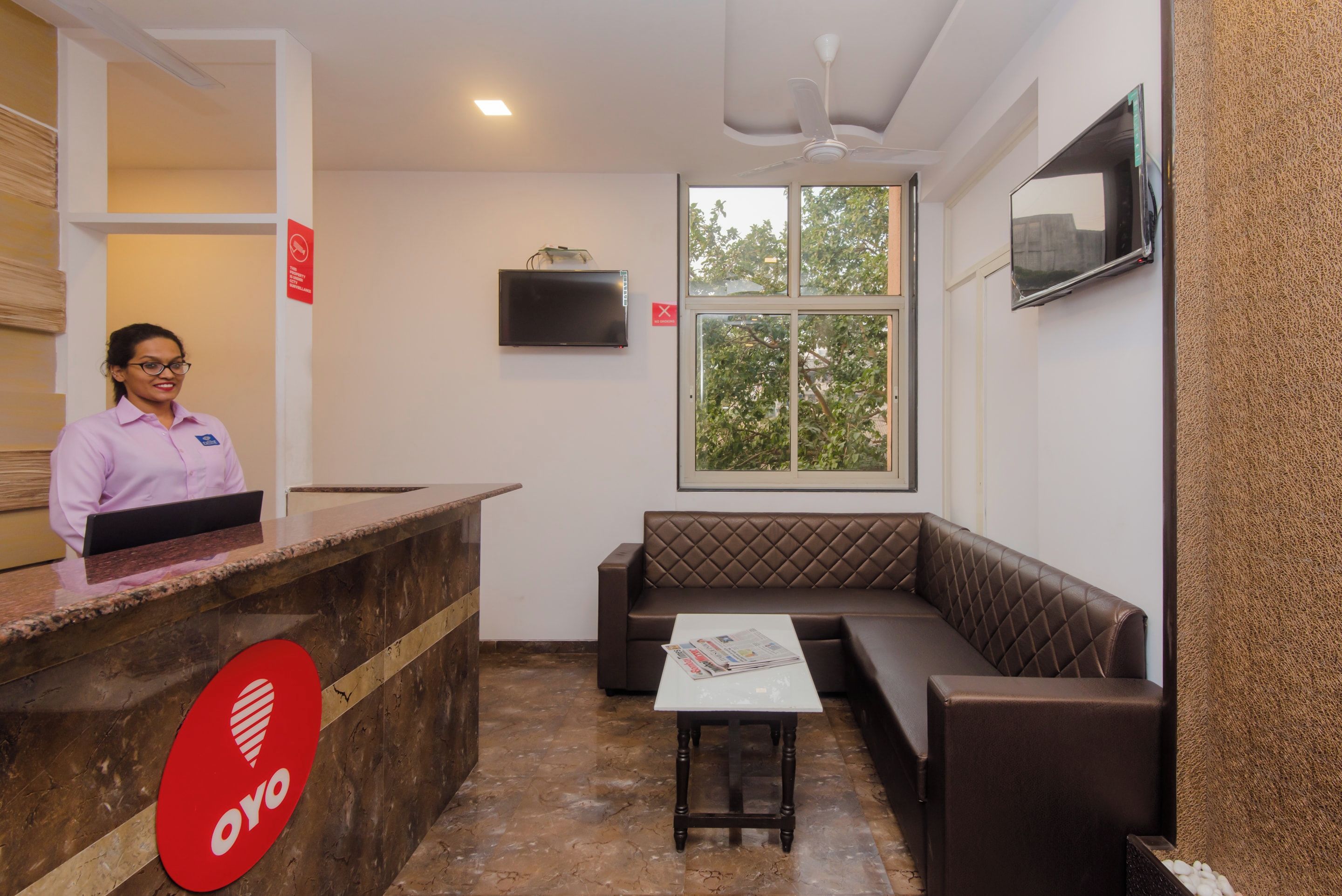 A reception area with a receptionist, a reception desk, a seating area with a bench, a television mounted on the wall, and a window with a view of trees.