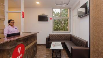 A reception area with a receptionist, a reception desk, a seating area with a bench, a television mounted on the wall, and a window with a view of trees.