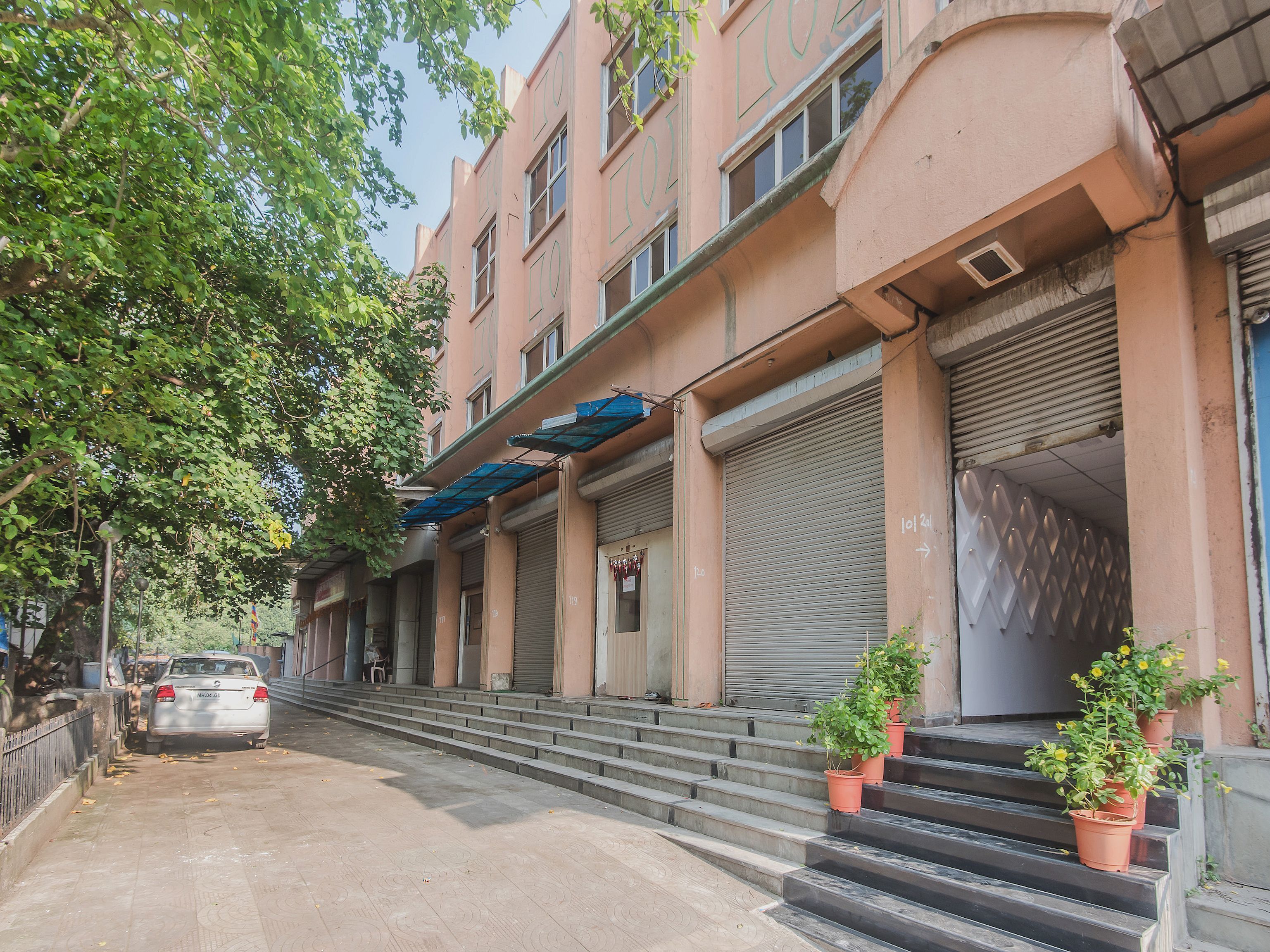 A multi-story building with closed shutters, a car parked in front, and potted plants on the steps.