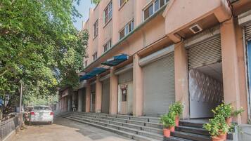 A multi-story building with closed shutters, a car parked in front, and potted plants on the steps.
