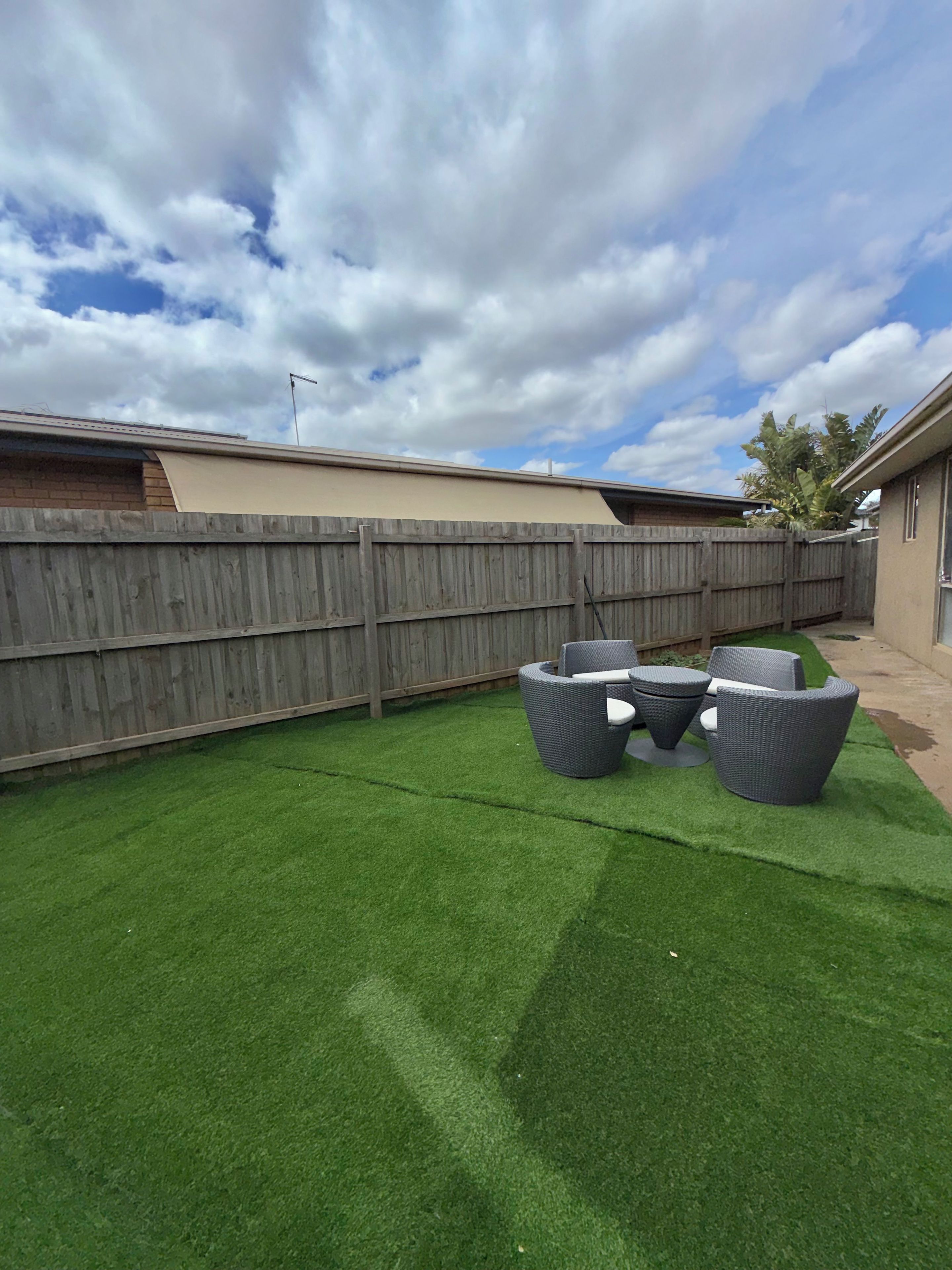 Un patio con césped artificial, cerca de madera y mobiliario de mimbre.