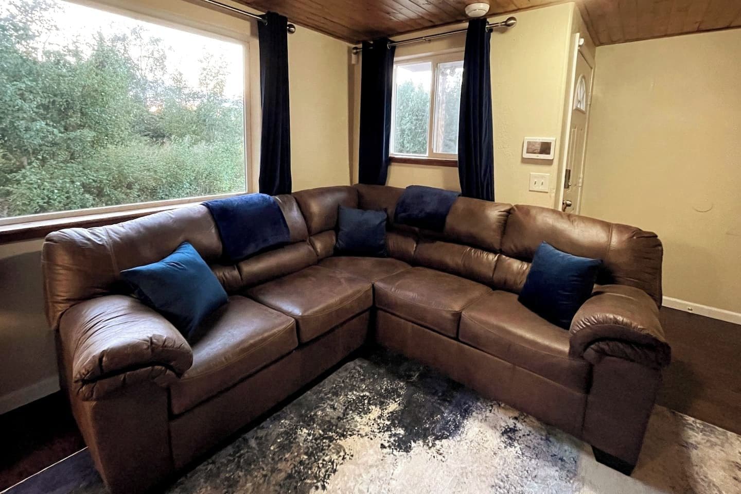 A brown leather sectional sofa with blue cushions, positioned in a room with a large window and a view of greenery outside.