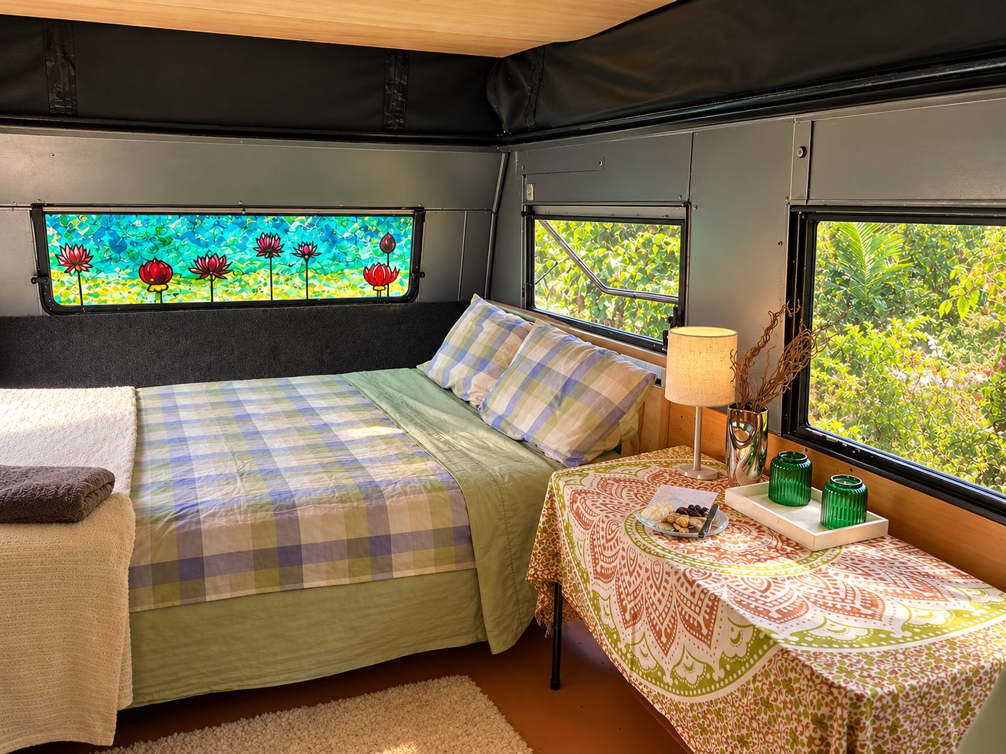 A cozy interior with a bed, a table with a lamp, and a window with a view of greenery.