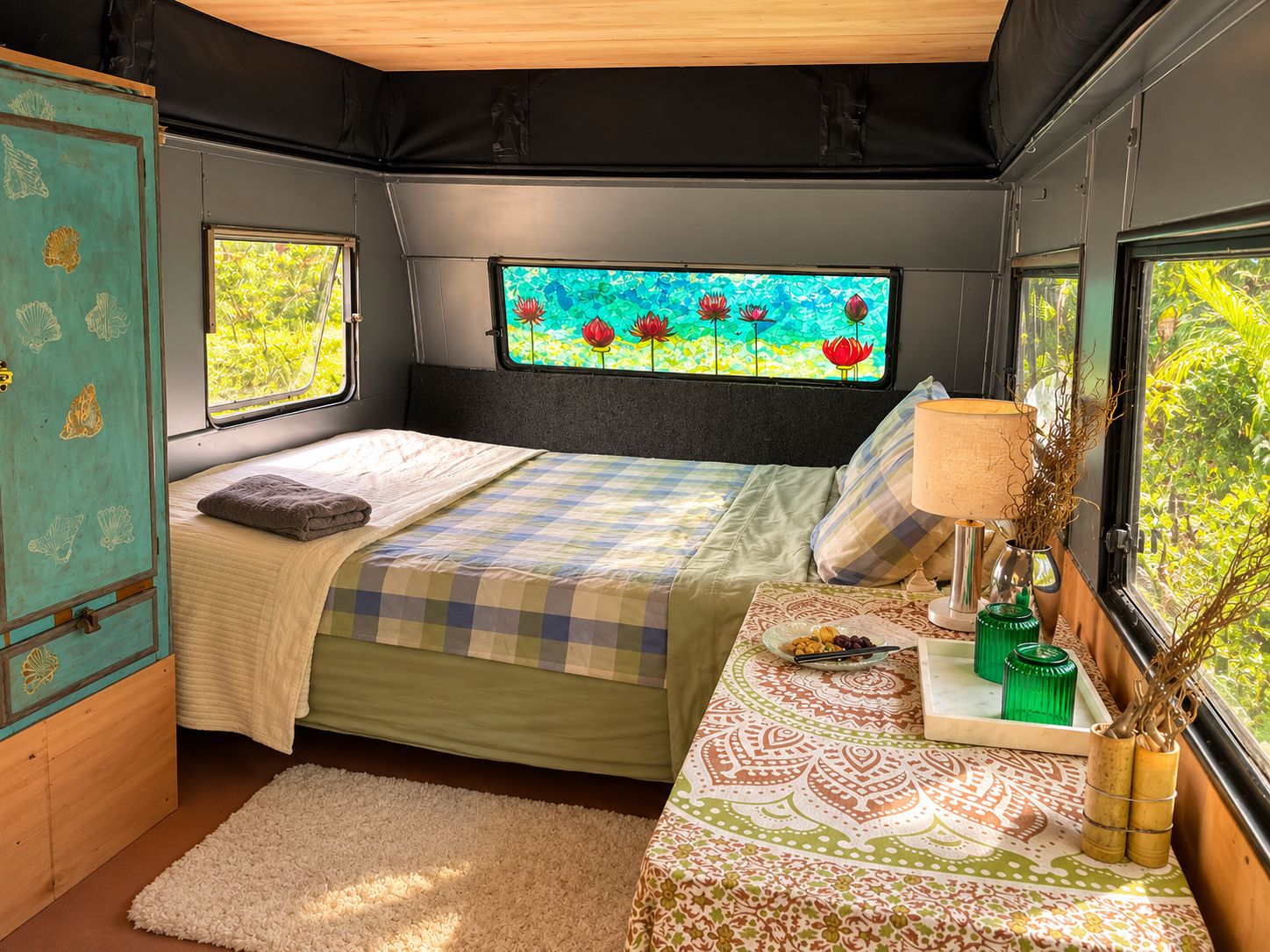 Une chambre à coucher de taille modeste, comprenant un lit, une fenêtre donnant sur un jardin fleuri, une table de chevet avec une lampe et un petit meuble de rangement.