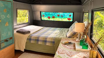 Une chambre à coucher de taille modeste, comprenant un lit, une fenêtre donnant sur un jardin fleuri, une table de chevet avec une lampe et un petit meuble de rangement.