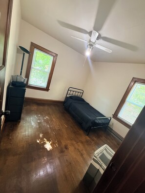 Une chambre avec un lit, un bureau, un ventilateur de plafond et des fenêtres équipées de stores.