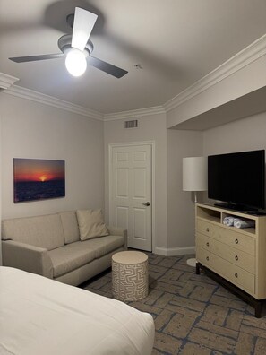 Una habitación de hotel con cama, sofá, televisor, ventilador de techo, un cuadro y una puerta.