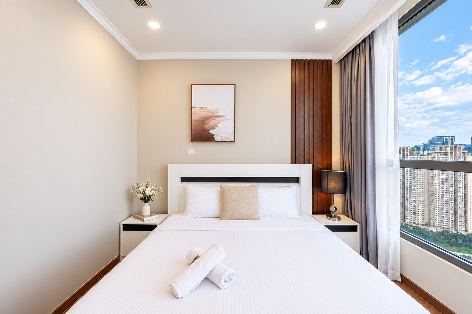 A hotel room with a large bed, a nightstand with a lamp, a painting on the wall, and a view of the city through the window.