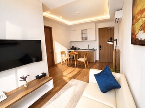 A modern living room with a flat-screen TV, a sofa with a blue pillow, a wooden coffee table, and a kitchen in the background. - One Studio Suite (Poipet)