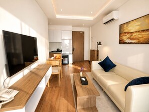 A modern living room with a flat-screen TV, a wooden coffee table, a beige sofa with a blue pillow, and a kitchen in the background. - One Studio Suite (Poipet)