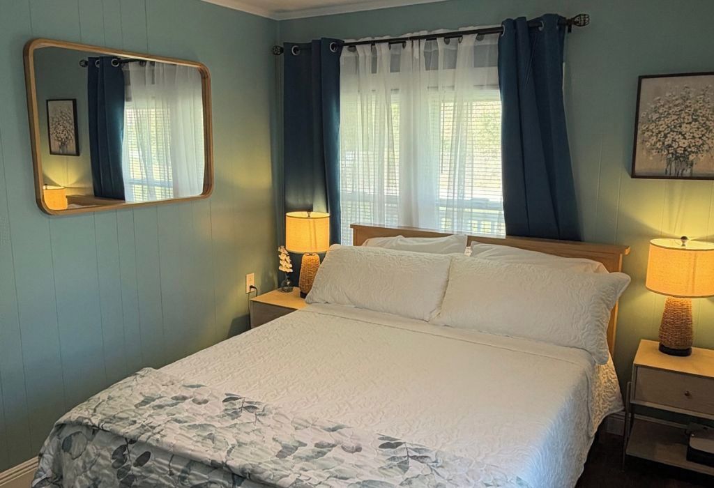 Een slaapkamer met een bed, nachtkastje, lamp, spiegel en gordijnen.
